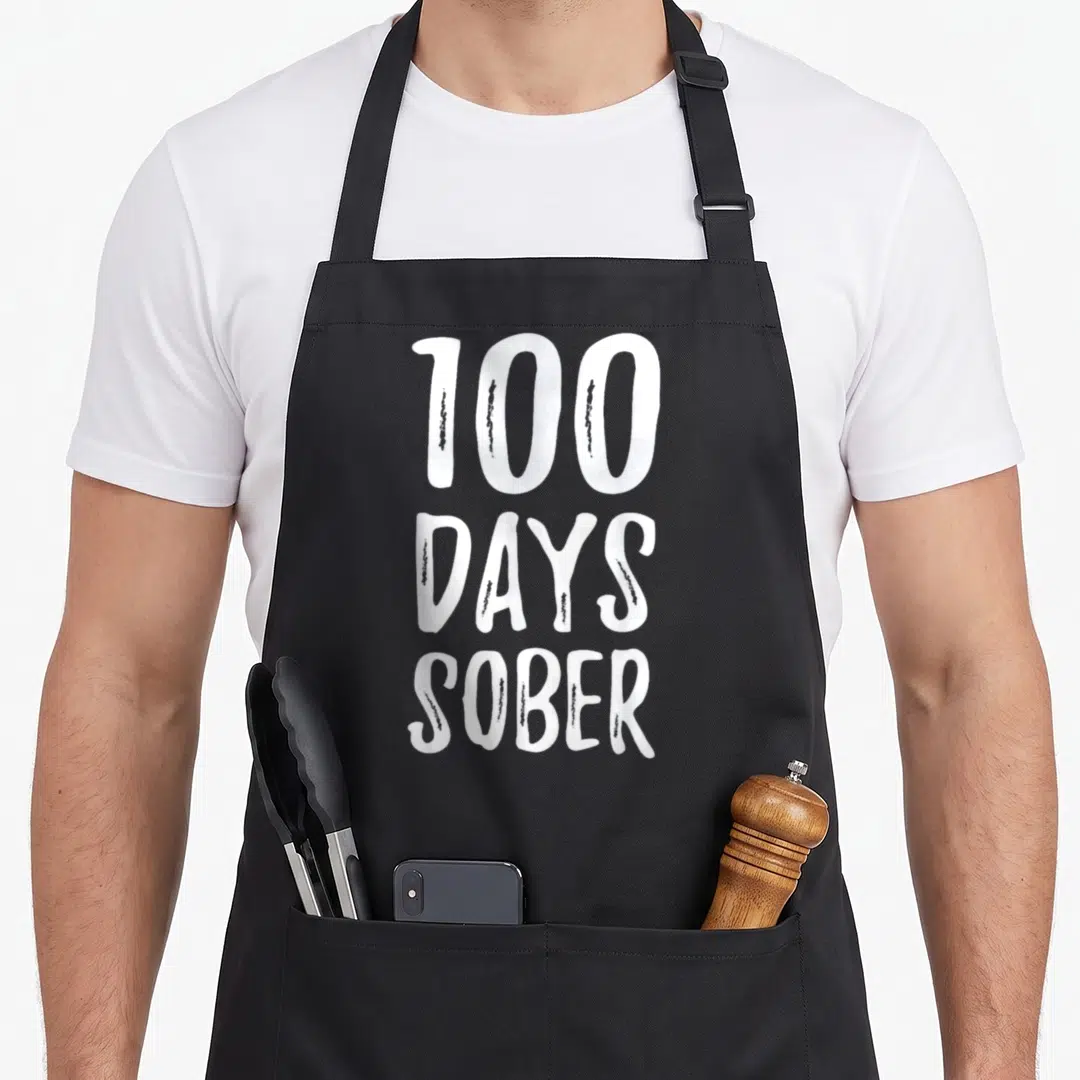 Sober congratulations apron - White 100 DAYS SOBER stacked typography on grey background — shown on a black cotton canvas apron against a white background