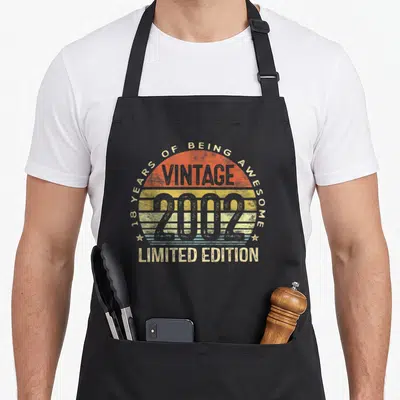 Edition 18th birthday apron - Striped sunset circle with 18 years of being awesome vintage 2002 limited edition text — shown on a black cotton canvas apron against a white background