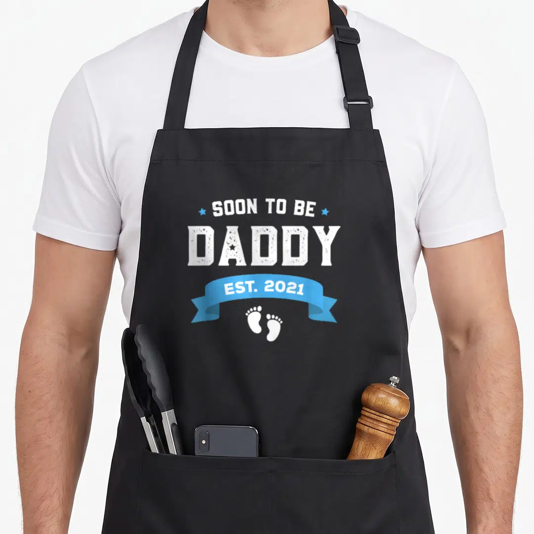 Cute soon daddy apron - SOON TO BE DADDY EST 2021 text with blue ribbon and baby footprints — shown on a black cotton canvas apron against a white background