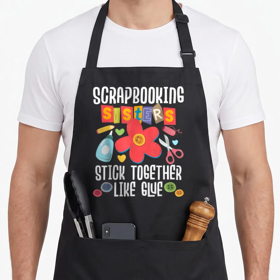 Scrapbooking diying apron - Scrapbooking sisters stick together like glue text with red flower and craft supplies — shown on a black cotton canvas apron against a white background