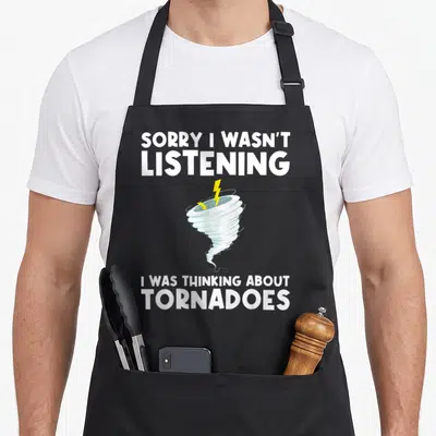Tornado hurricane apron - Cartoon tornado with yellow lightning bolt framed by white text saying sorry I wasn't — shown on a black cotton canvas apron against a white background
