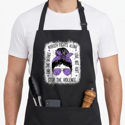 Stop domestic aprons - Cartoon woman head with purple bow and sunglasses surrounded by circular text about stopping — shown on a black cotton canvas apron against a white background
