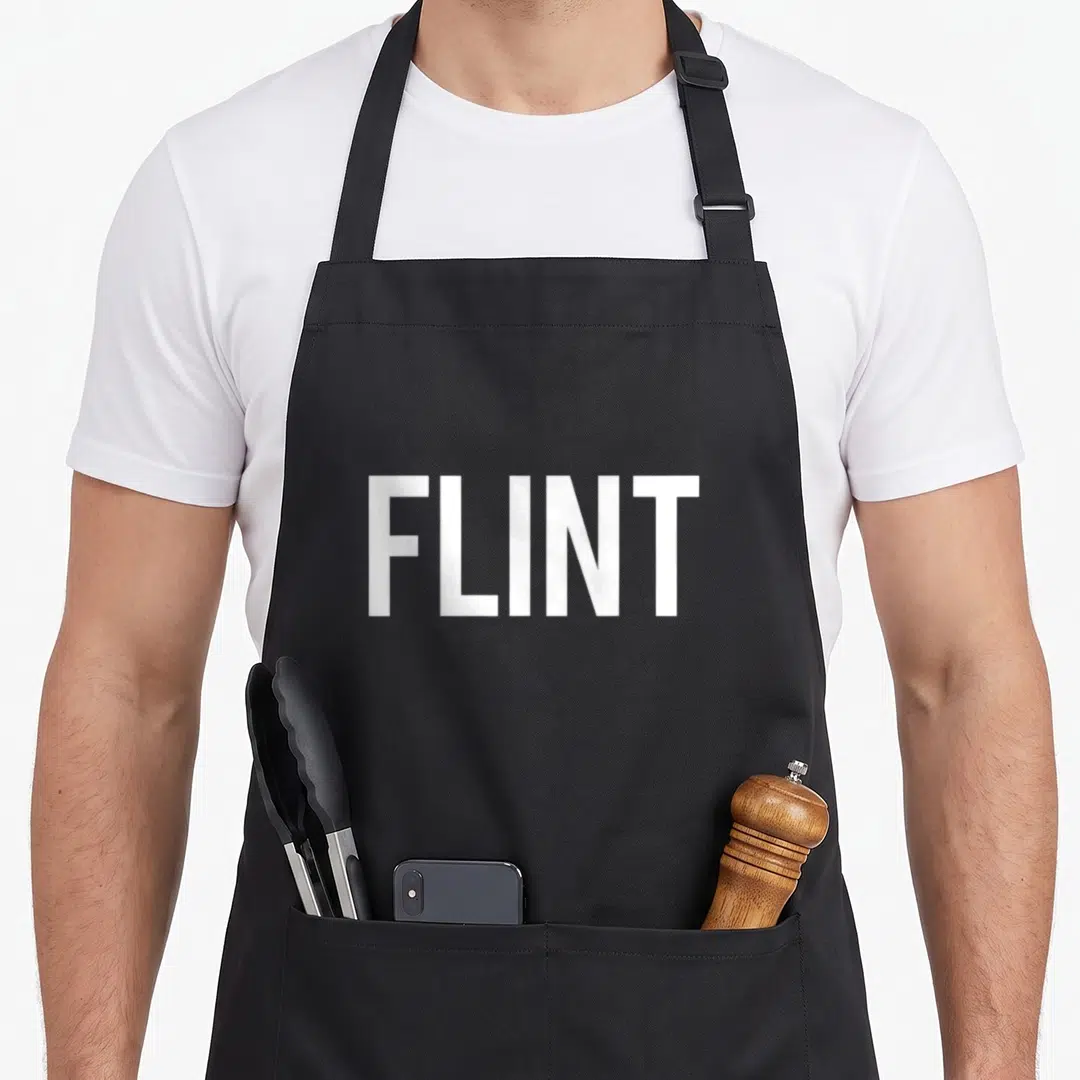 Flint michigan apron - White block text FLINT printed on a black background — shown on a black cotton canvas apron against a white background