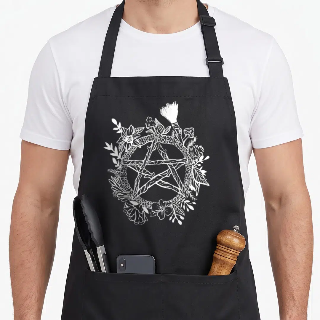 Pentacle goth apron - White line art pentagram wreath with flowers mushrooms and broom on black background — shown on a black cotton canvas apron against a white background