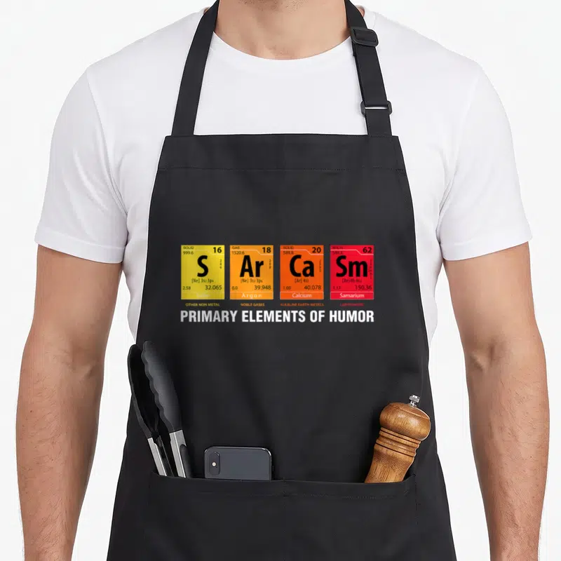 Sarcasm element table apron - Four periodic table boxes S Ar Ca Sm labeled PRIMARY ELEMENTS OF HUMOR — shown on a black cotton canvas apron against a white background