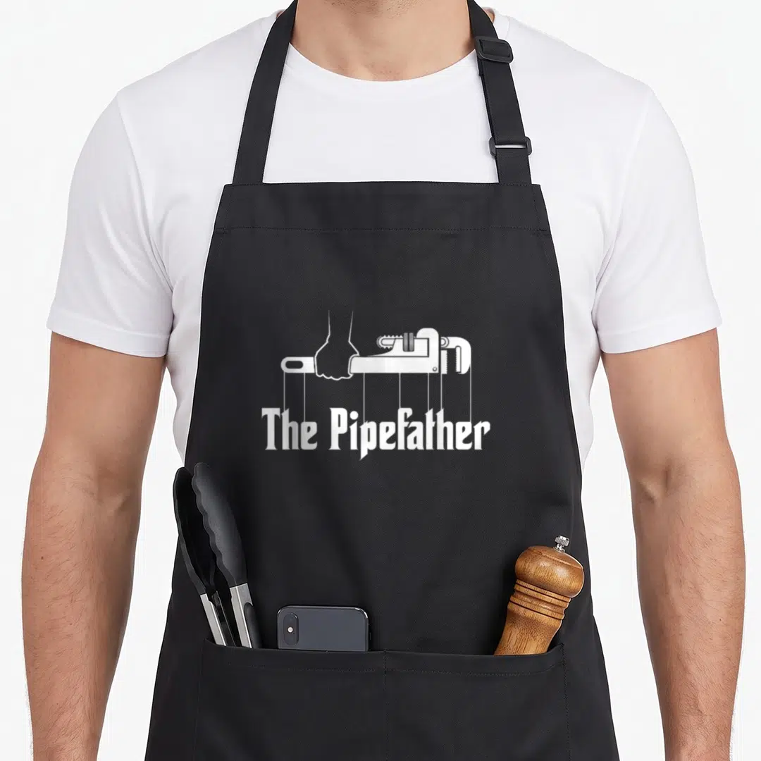 Pipefather plumber apron - White hand holding pipe wrench with vertical lines connecting to text The Pipefather — shown on a black cotton canvas apron against a white background