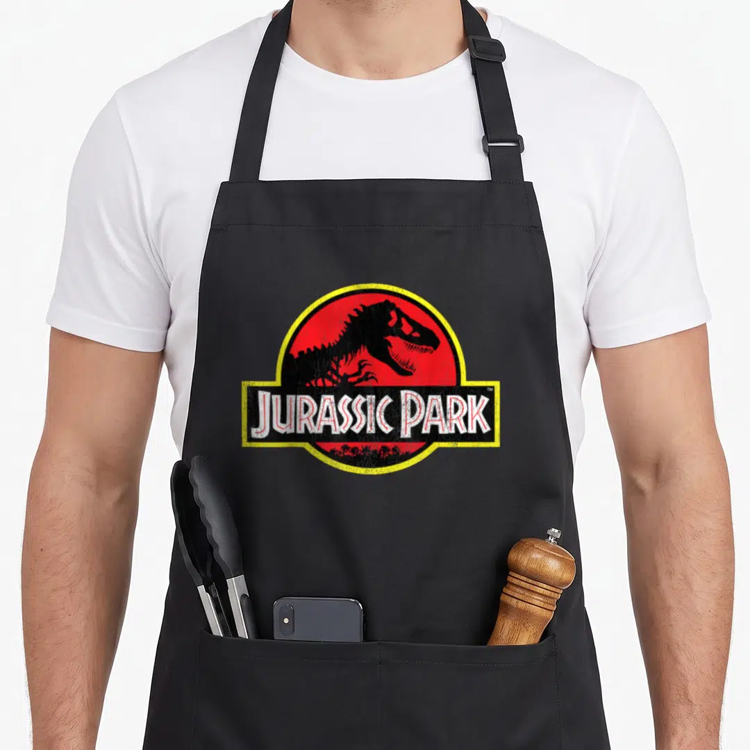 Distressed crew apron - Black dinosaur skeleton inside red circle with yellow border and JURASSIC PARK text — shown on a black cotton canvas apron against a white background