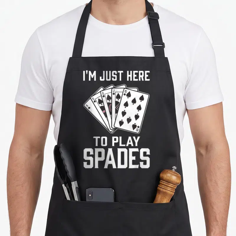 Spade game aprons - I'M JUST HERE TO PLAY SPADES text surrounding five fanned playing cards in white, black, and pink — shown on a black cotton canvas apron against a white background