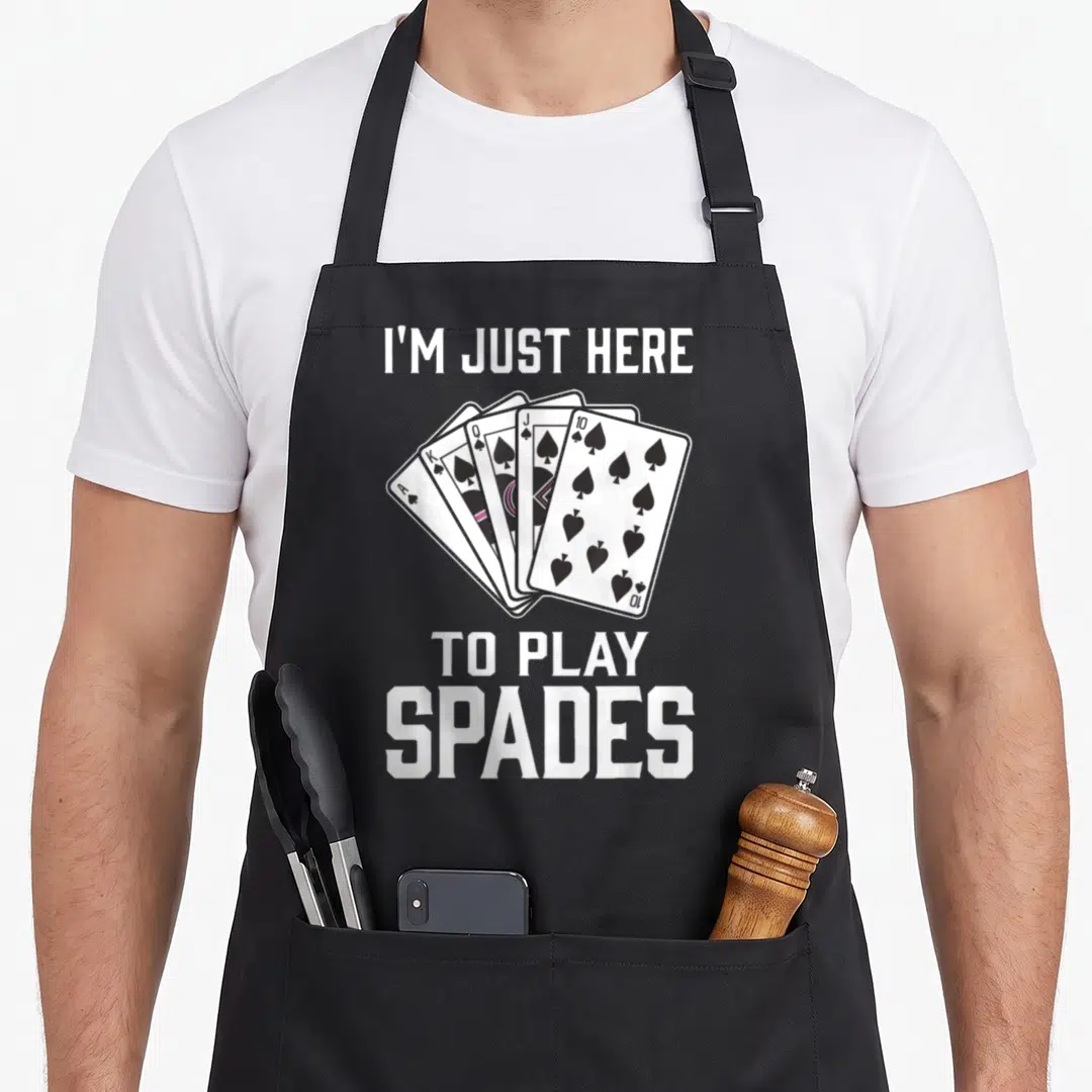 Spade game aprons - I'M JUST HERE TO PLAY SPADES text surrounding five fanned playing cards in white, black, and pink — shown on a black cotton canvas apron against a white background