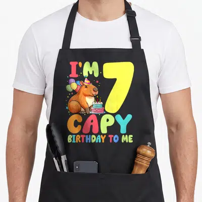 Capy 7th birthday apron - Cartoon capybara with balloons and cake next to number 7 and colorful text — shown on a black cotton canvas apron against a white background