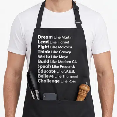 Dream novelty apron - Ten lines of white text listing verbs like Dream, Lead, Fight paired with names like Martin — shown on a black cotton canvas apron against a white background