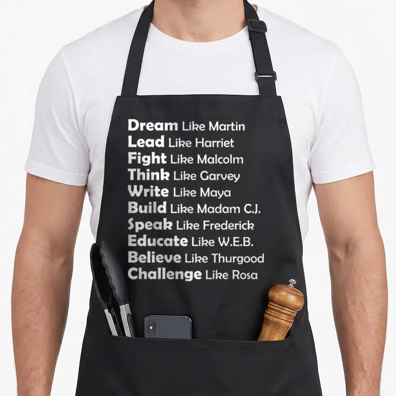 Dream novelty apron - Ten lines of white text listing verbs like Dream, Lead, Fight paired with names like Martin — shown on a black cotton canvas apron against a white background