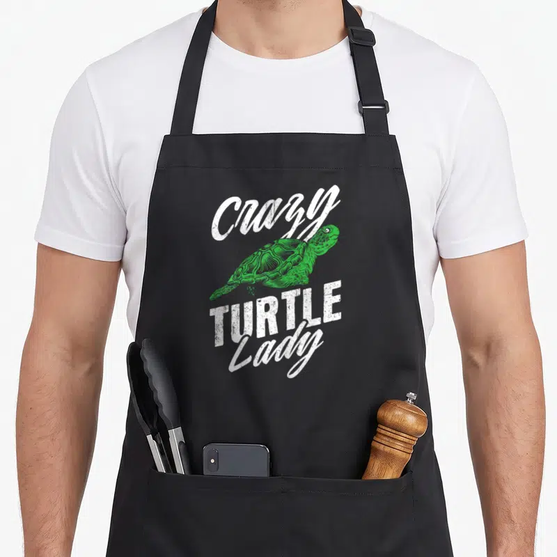 Retro turtle apron - Green turtle illustration surrounded by distressed white Crazy TURTLE Lady text on black background — shown on a black cotton canvas apron against a white background