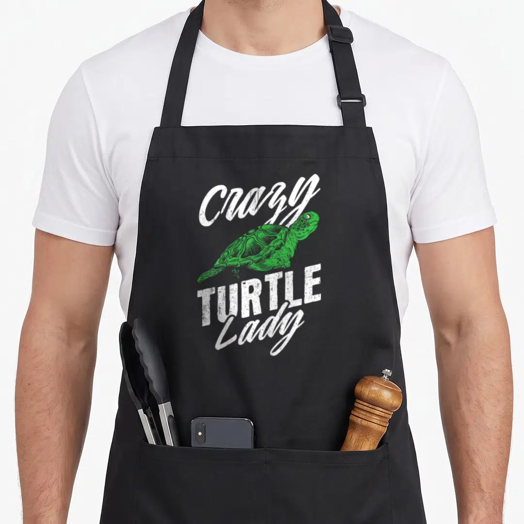 Retro turtle apron - Green turtle illustration surrounded by distressed white Crazy TURTLE Lady text on black background — shown on a black cotton canvas apron against a white background