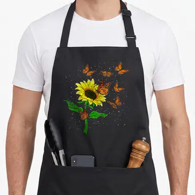 Blossom sunflower apron - Yellow sunflower with green stem and six orange butterflies in watercolor style on black — shown on a black cotton canvas apron against a white background