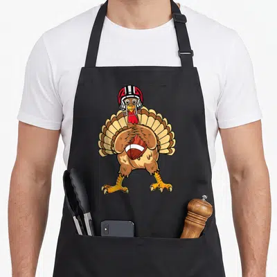 Playing thanksgiving apron - Cartoon turkey in football helmet holding ball with brown red and yellow coloring — shown on a black cotton canvas apron against a white background
