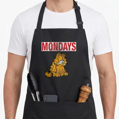 Monday cat apron - Red MONDAYS text above grumpy orange cat in distressed style — shown on a black cotton canvas apron against a white background