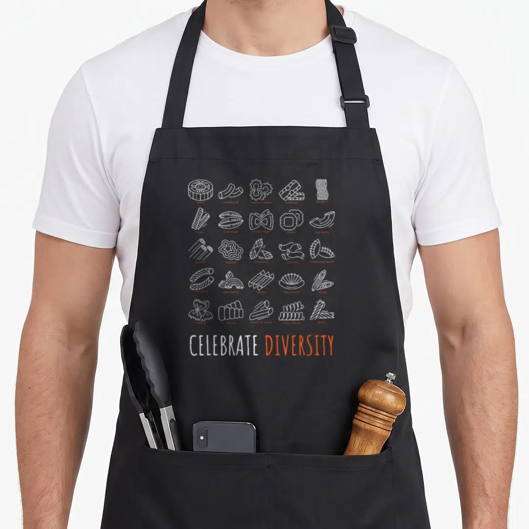 Celebrate pasta apron - Grid of white line-art pasta shapes and CELEBRATE DIVERSITY text in white and orange — shown on a black cotton canvas apron against a white background