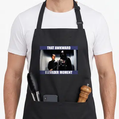 Elevader meme apron - Photo of two characters between blue bars with text That Awkward Elevader Moment — shown on a black cotton canvas apron against a white background