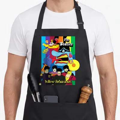 Collage beatle apron - Cartoon Beatles standing below a blue monster and yellow submarine against rainbow stripes — shown on a black cotton canvas apron against a white background