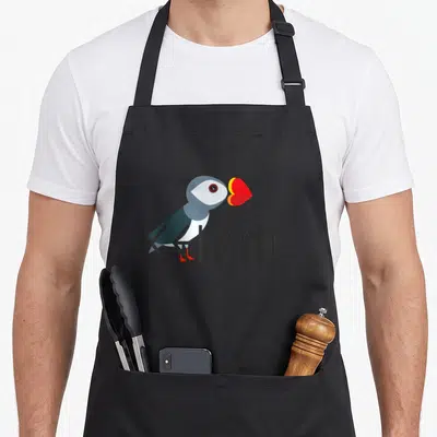 Puffin icelandic apron - Stylized puffin in grey white red and yellow standing over black ICELAND text — shown on a black cotton canvas apron against a white background