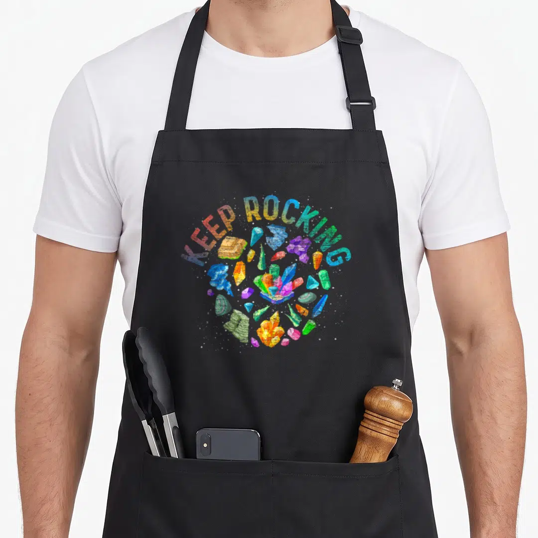 Rock colorful aprons - KEEP ROCKING text arched over blue purple green yellow orange red crystals — shown on a black cotton canvas apron against a white background