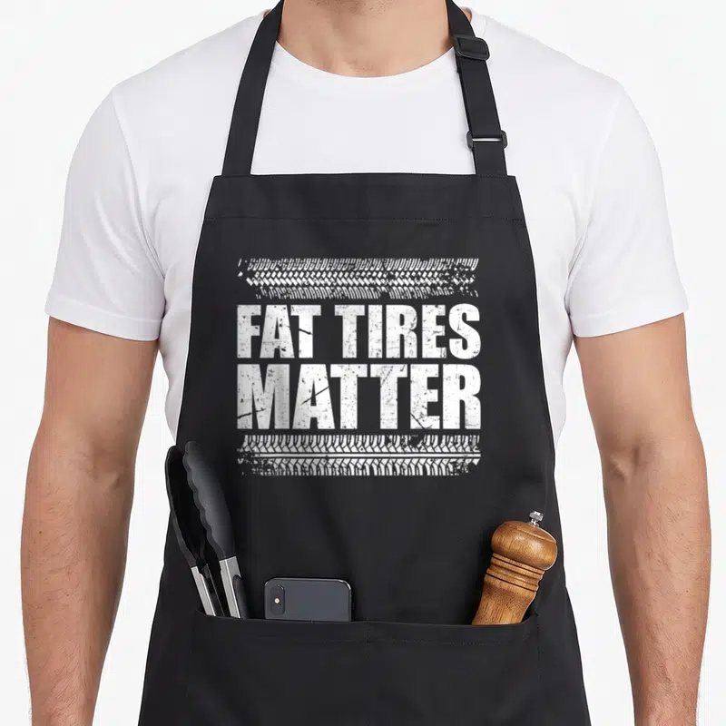 Drag racing car apron - White fat tires matter text with tire tread patterns above and below — shown on a black cotton canvas apron against a white background