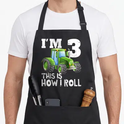 3rd farm apron - Green cartoon tractor with I'm 3 This Is How I Roll text — shown on a black cotton canvas apron against a white background
