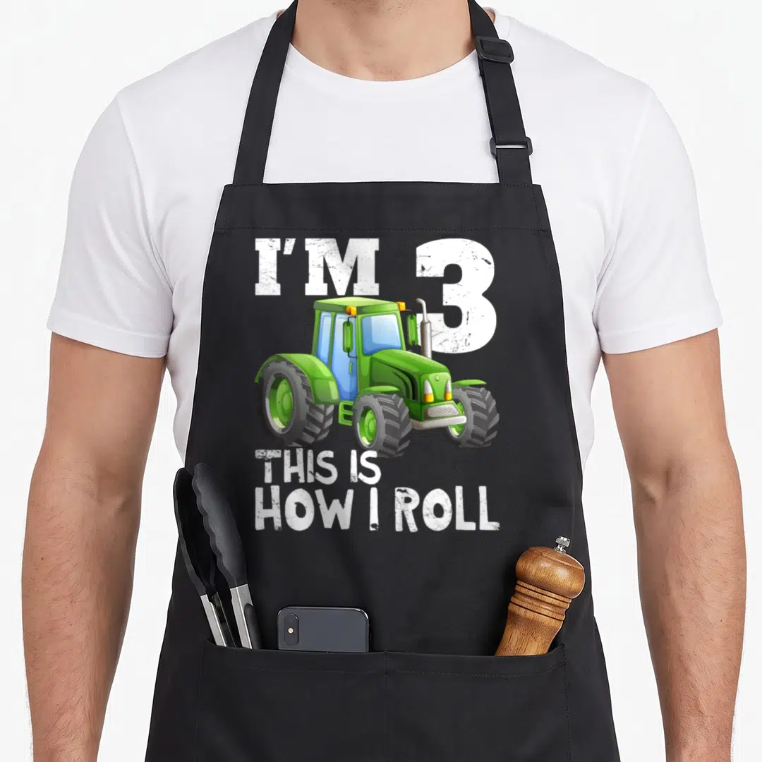 3rd farm apron - Green cartoon tractor with I'm 3 This Is How I Roll text — shown on a black cotton canvas apron against a white background