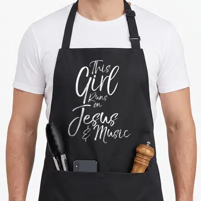 Musician christian apron - White stacked script text reading This Girl Runs on Jesus and Music — shown on a black cotton canvas apron against a white background