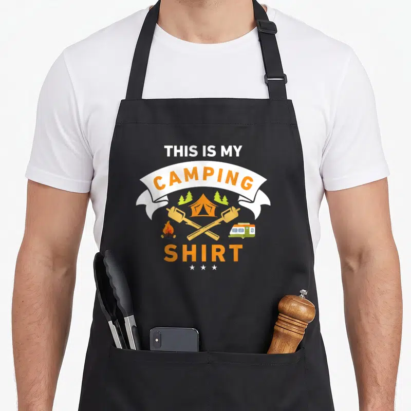 Camper camping apron - THIS IS MY CAMPING SHIRT text with tent campfire van marshmallows and three stars in orange — shown on a black cotton canvas apron against a white background
