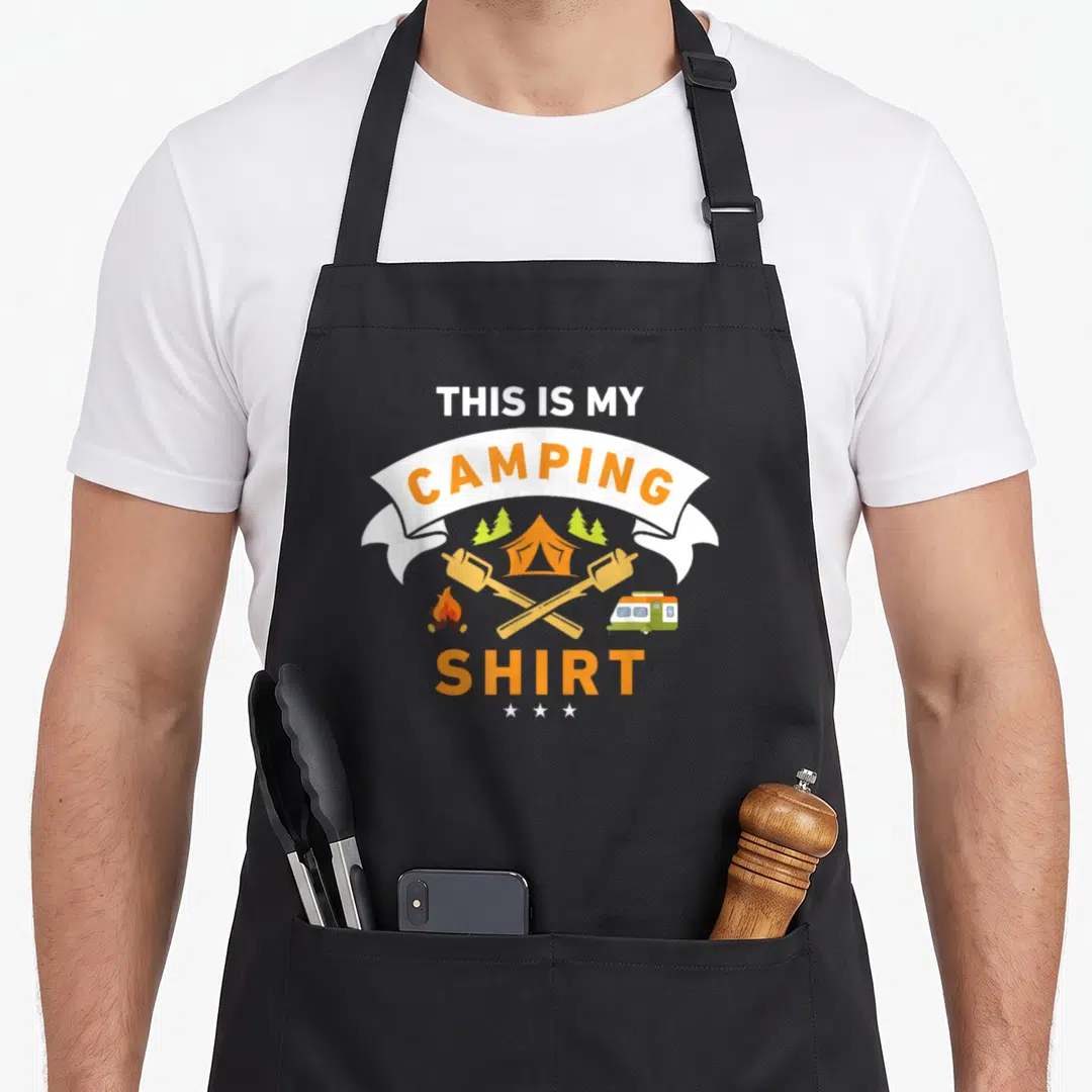 Camper camping apron - THIS IS MY CAMPING SHIRT text with tent campfire van marshmallows and three stars in orange — shown on a black cotton canvas apron against a white background