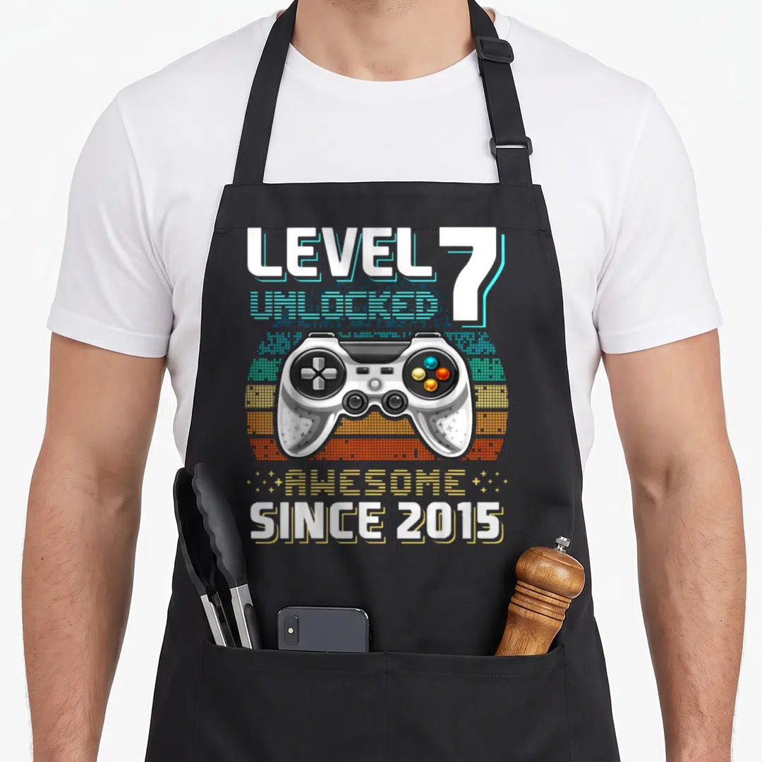 7th birthday video game apron - Retro game controller over pixelated sun with Level 7 Unlocked text in blue and yellow — shown on a black cotton canvas apron against a white background