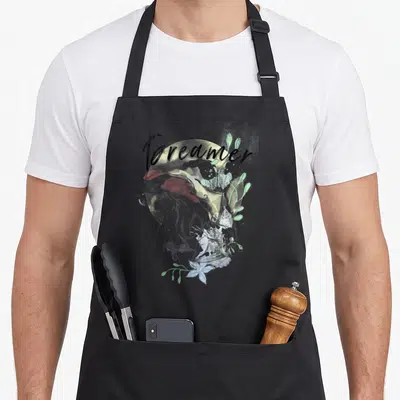 Ariel paint apron - Mermaid resting chin on hands with Dreamer text and watercolor plants in grey, green, blue, and red — shown on a black cotton canvas apron against a white background