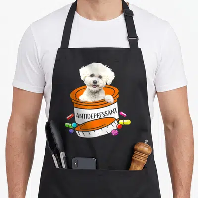 Antidepressant bichon frise apron - White dog in orange pill bottle labeled ANTIDEPRESSANT with scattered colorful — shown on a black cotton canvas apron against a white background