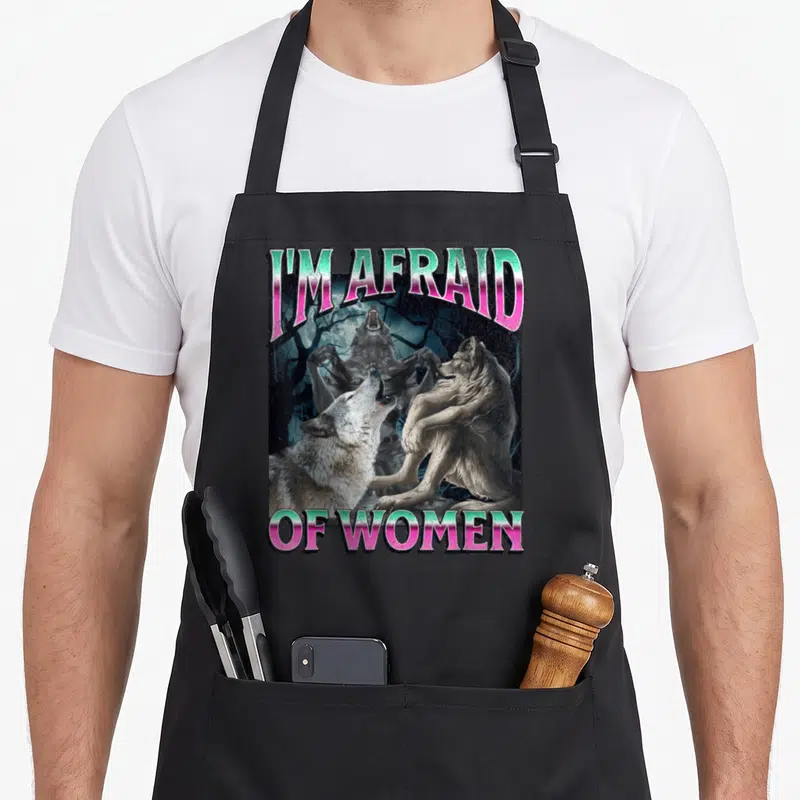 Afraid meme apron - I'M AFRAID OF WOMEN text with werewolves and moon in pink green grey black — shown on a black cotton canvas apron against a white background