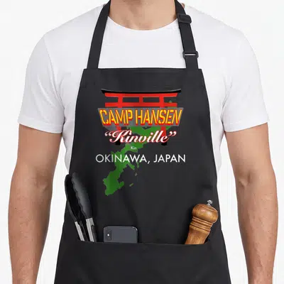 Camp japan apron - Red torii gate over green map with Camp Hansen Kinville Okinawa text — shown on a black cotton canvas apron against a white background