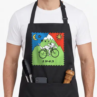 Lsd bicycle apron - Stamp-style print showing person on bike, moon, sun, mountain, and text 25 1943 in blue red green — shown on a black cotton canvas apron against a white background
