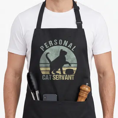 Cat personal printed aprons - Retro circle with cat silhouette and bowl, text reading PERSONAL CAT SERVANT in teal — shown on a black cotton canvas apron against a white background