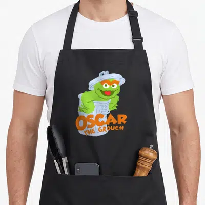 Flat oscar the grouch apron - Green Oscar monster smiling in light blue bin with orange OSCAR THE GROUCH text — shown on a black cotton canvas apron against a white background