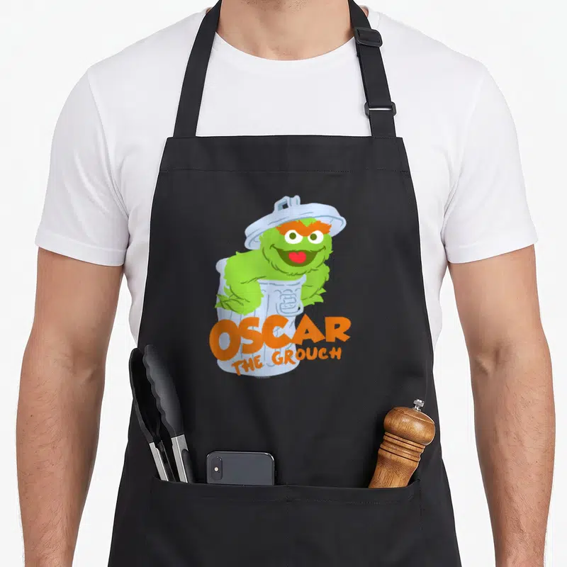 Flat oscar the grouch apron - Green Oscar monster smiling in light blue bin with orange OSCAR THE GROUCH text — shown on a black cotton canvas apron against a white background