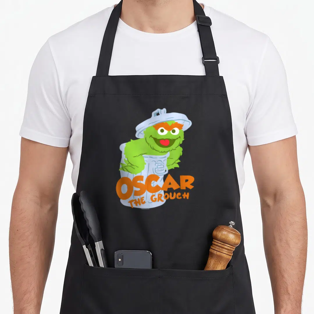 Flat oscar the grouch apron - Green Oscar monster smiling in light blue bin with orange OSCAR THE GROUCH text — shown on a black cotton canvas apron against a white background