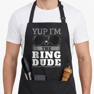 Yup wedding apron - YUP I'M THE RING DUDE text with black sunglasses and diamond ring graphic — shown on a black cotton canvas apron against a white background