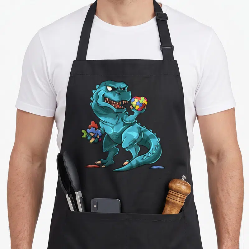 Puzzle dinosaur apron - Teal cartoon dinosaur holding a colorful puzzle piece heart and loose jigsaw fragments — shown on a black cotton canvas apron against a white background