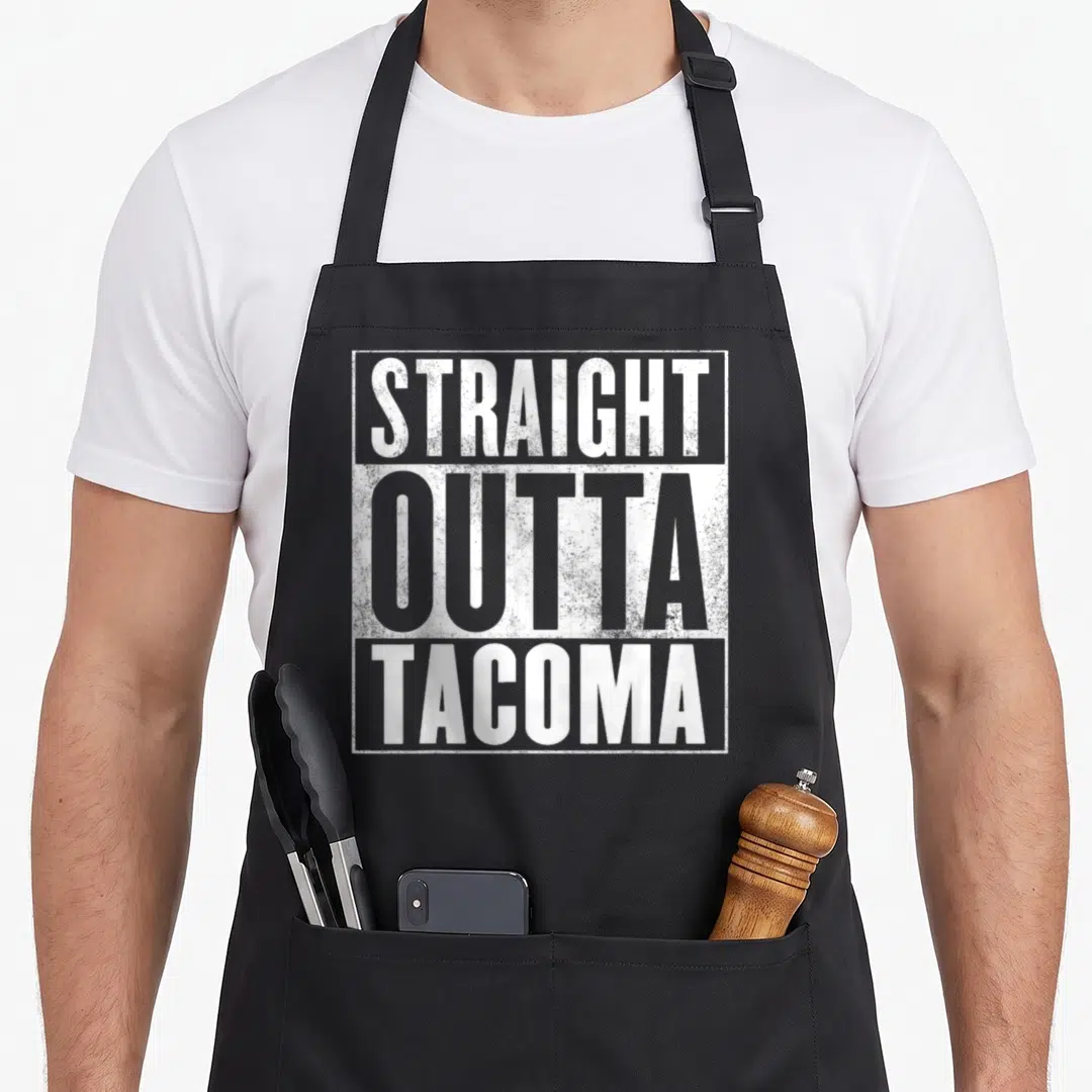 Straight tacoma apron - Stacked white and grey text reading STRAIGHT OUTTA TACOMA inside a distressed rectangular border — shown on a black cotton canvas apron against a white background
