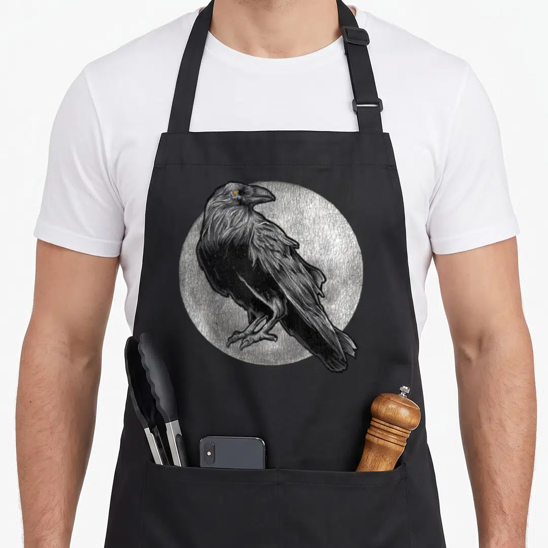 Moon bird apron - Raven illustration in front of a textured white moon circle with orange eye detail — shown on a black cotton canvas apron against a white background