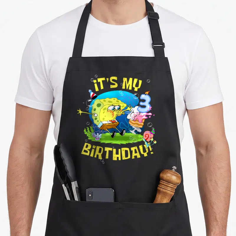 3rd cake apron - SpongeBob blowing out a number 3 candle with IT'S MY BIRTHDAY! text and Gary wearing a party hat — shown on a black cotton canvas apron against a white background