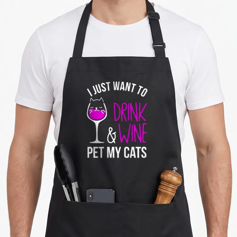 Wine cat apron - I Just Want To Drink Wine Pet My Cats text around magenta cat in wine glass outline — shown on a black cotton canvas apron against a white background