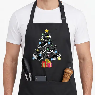 Orca christmas tree apron - Cartoon orcas form a Christmas tree shape with a yellow star and three wrapped gifts — shown on a black cotton canvas apron against a white background