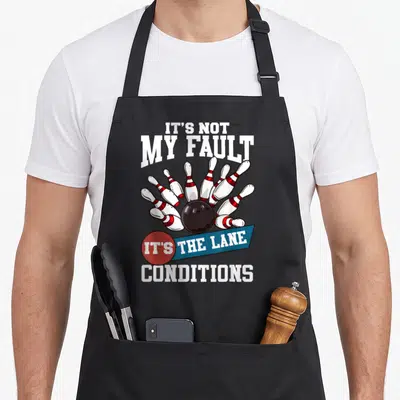 Fault bowling life apron - Cartoon bowling ball hitting pins with It's Not My Fault text — shown on a black cotton canvas apron against a white background